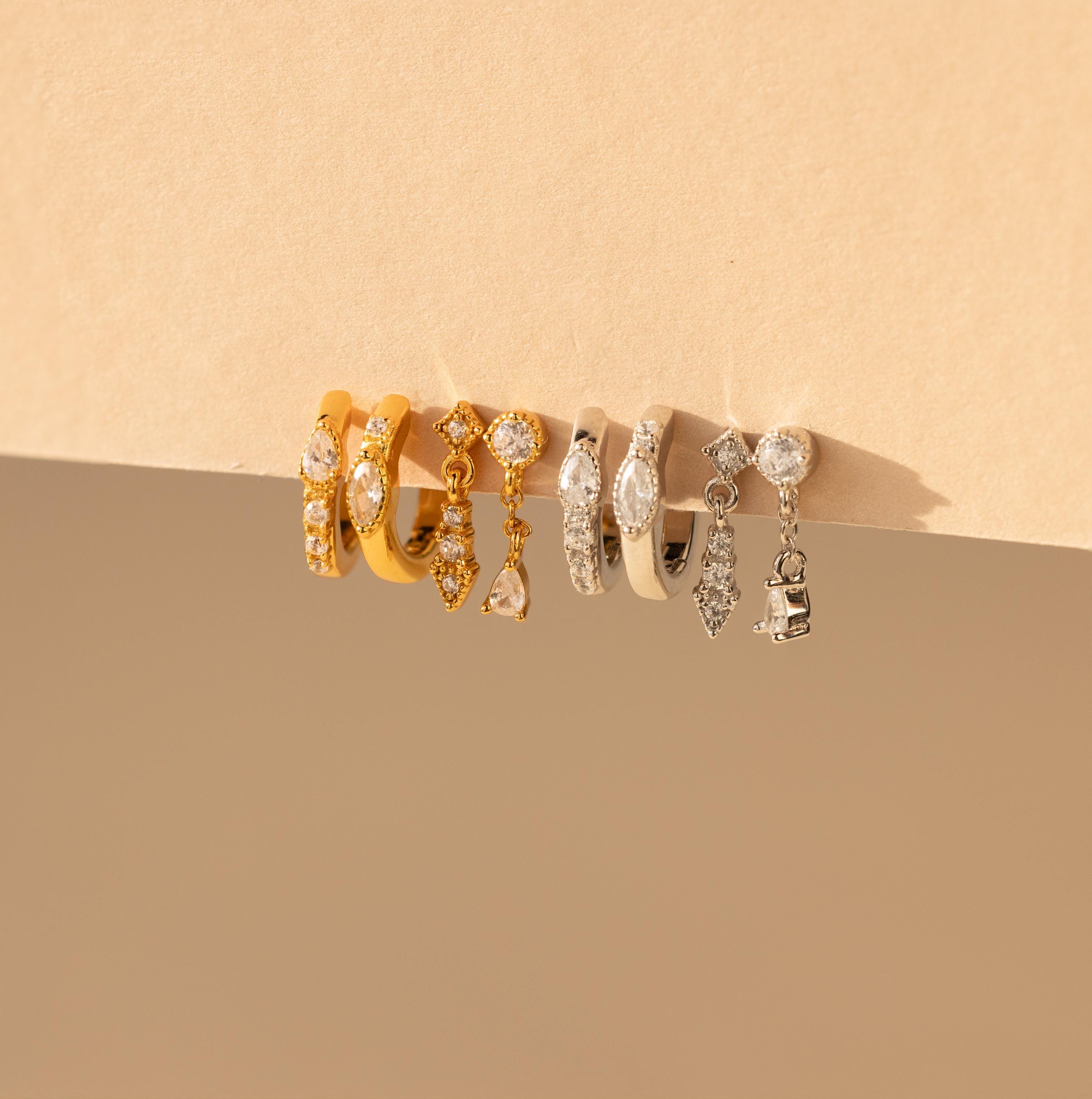 A set of earrings displayed against a neutral background, including styles with pave stones and charms in both gold and silver finishes.